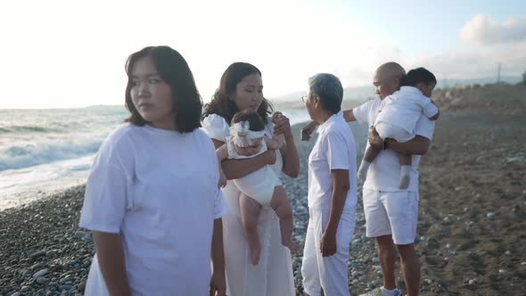 Thoughtful Young Asian Woman Holding Baby Girl with Family Around at Mediterranean Sea Shore alt