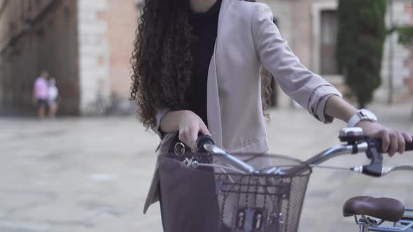 Pretty Woman With Long And Curly Hair Is Walking Along The Street With Bicycle alt