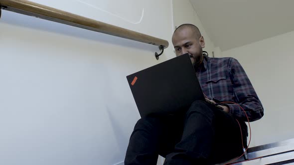 An anxious Asian office employee working on his laptop computer sitting on the steps of a staircase, alt