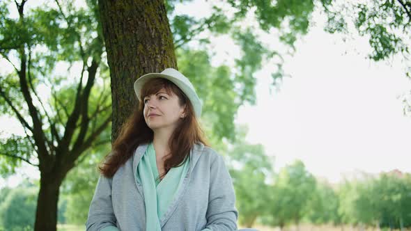 Happy Woman with a Hat Sits in the Park Near the Tree and Looks Around alt