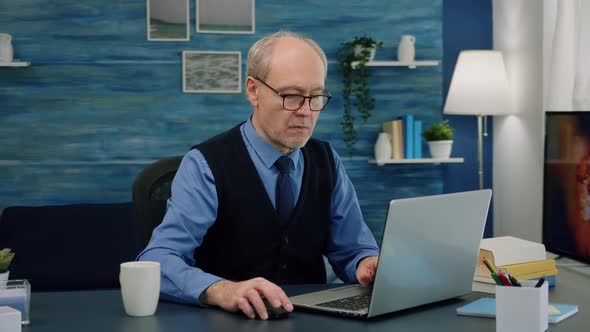 Tired Elderly Man Sitting at Desk Drinking Coffee Reading and Typing at Laptop alt
