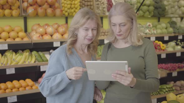 Portrait of Positive Caucasian Senior Women Using Tablet in Grocery. Two Housewives Standing Between alt