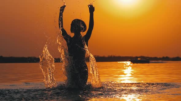 Silhouette of Woman at Sunset Raises Hands Up and Creating Splashes of Water. Slow Motion alt