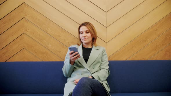 Happy Young Woman Using Smartphone While Sitting on a Sofa at Lobby Hotel alt