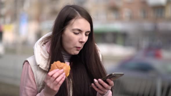Happy Woman Eating a Burger Holding a Smart Phone Walking Outdoors in the City