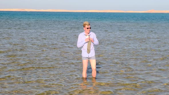 A Funny Man in a Shirt Shorts Socks and Sunglasses Adjusts His Tie Standing in the Sea Holding a