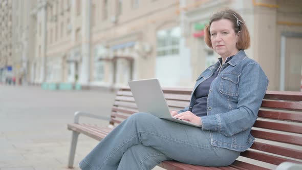Old Woman with Laptop Smiling at Camera While Sitting Outdoor on Bench alt