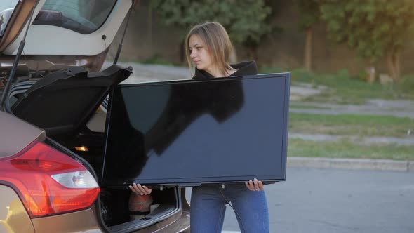 Woman Funny Trying to Put Purchased Modern Tv in Car Trunk at Store Parking in Closeup alt