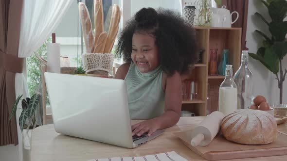 Little adorable afro African American schoolgirl studying online with laptop, at table in kitchen