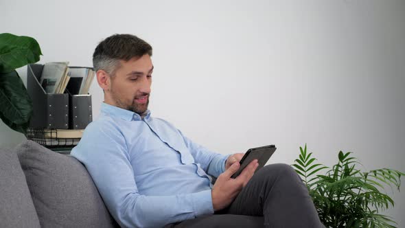 Smiling Businessman in a Light Blue Shirt Sitting on the Couch Using Tablet alt