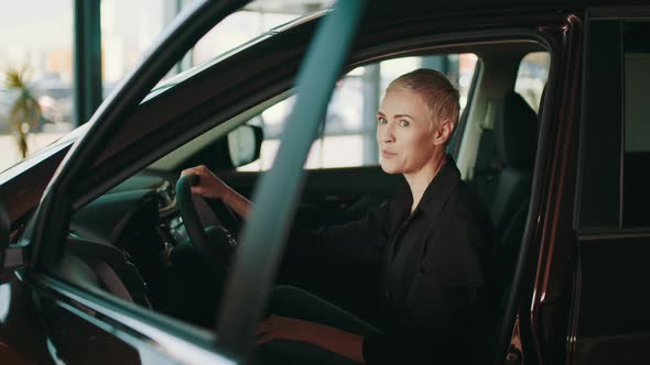 Cheerful Blond Woman Sitting in Modern Car with Open Door and Looking Aside alt