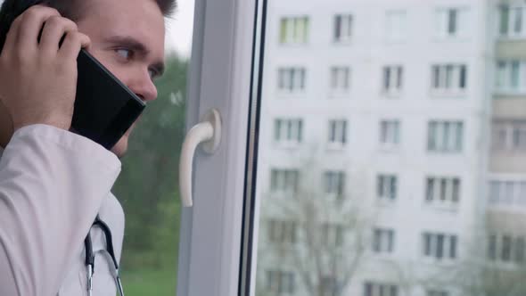 Doctor speaks on the phone near the window, against the background of a building