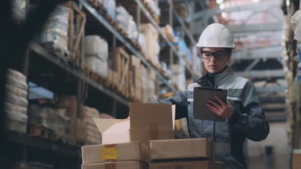 Woman in Warehouse in Helmet and Gray Uniform Opens a Cardboard Box