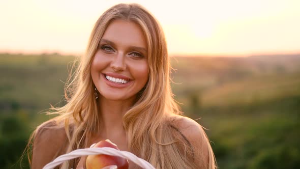 Beautiful sexy blonde girl in white dress posing in a field at sunset with a basket of fruit alt