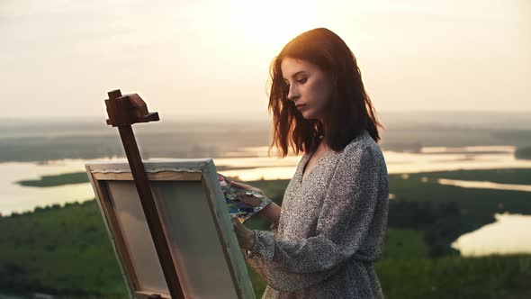 A Young Beautiful Woman Artist Draws a Painting on an Easel at Sunset Field alt