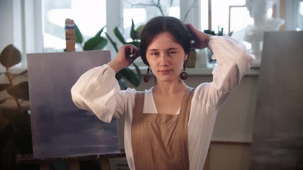 Young Woman Artist Lets Her Hair Out of Her Hairstyle and Smiles Holding a Brush alt
