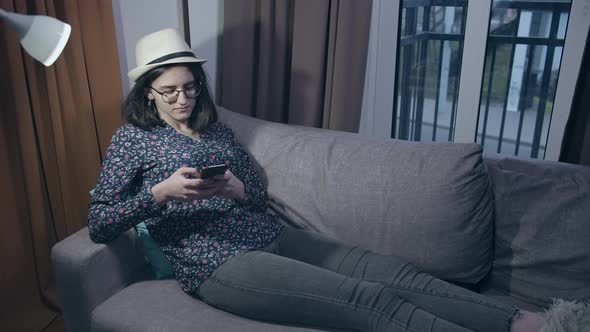 A Girl Student in Glasses and a Hat Reads Messages in Her Mobile Phone While Sitting on the Couch alt