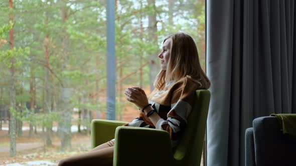 woman with a mug of tea sits in a chair by the window with a view of the forest