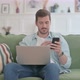 Young Man with Laptop Using Smartphone on Sofa
