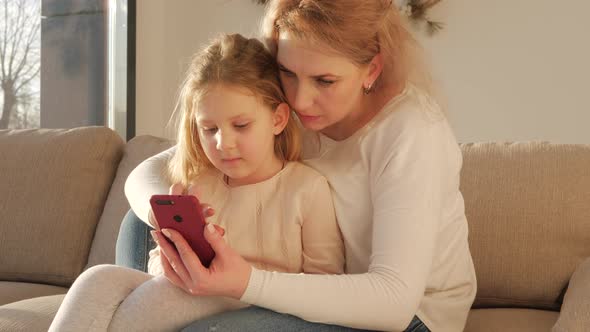 Cute little small kid child daughter having fun with adult mum enjoying using smart phone showing fu