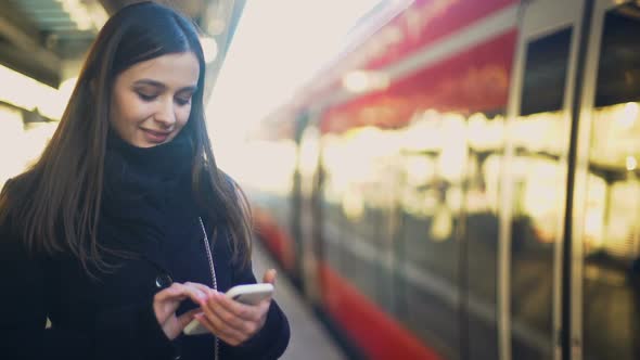 Young Lady Typing on Smartphone on Platform Near Train and Smiling to Camera alt