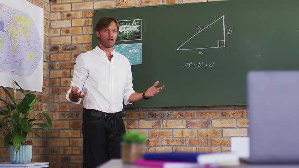 Caucasian male teacher teaching mathematics while looking at laptop in the classroom at school alt