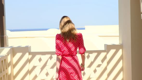 A Woman in a Red Dress Walks Down a Corridor Overlooking the Sea on a Sunny Summer Day