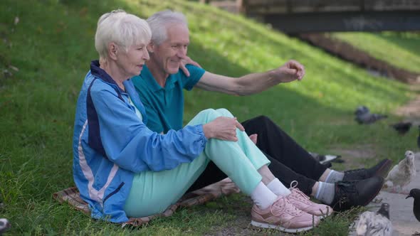 Loving Happy Senior Wife Hugging Husband Sitting on Green Summer Spring Meadow Feeding Birds in Slow alt