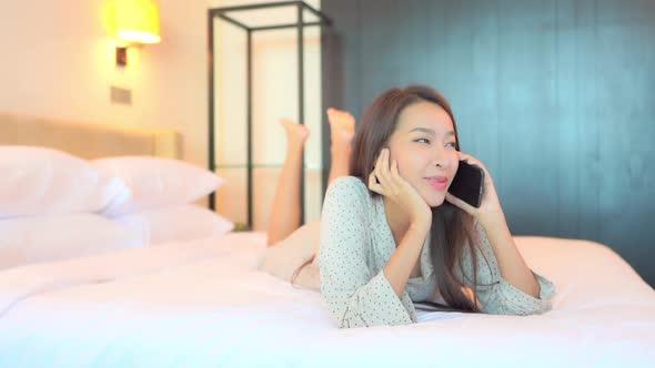 Smiling Young Asian Woman Talking on Smartphone With Friend or Lover in Privacy of Bedroom, Lying on alt