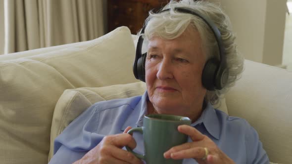Caucasian senior woman wearing headphones listening to music while holding coffee cup at home alt
