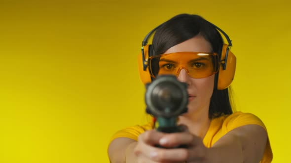 Brunette Woman in Protective Headphones and Glasses is Holding a Drill Like Gun alt