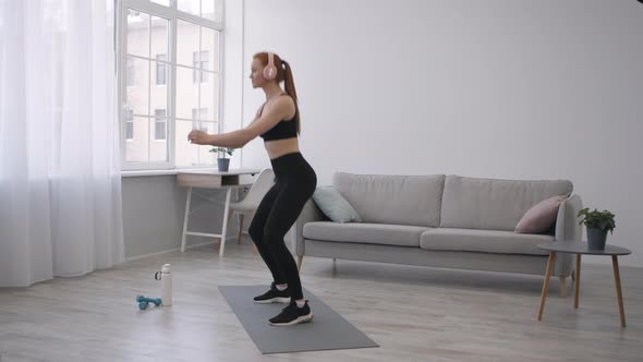 Athletic Woman Doing Jump Squats Exercising Wearing Headphones At Home alt
