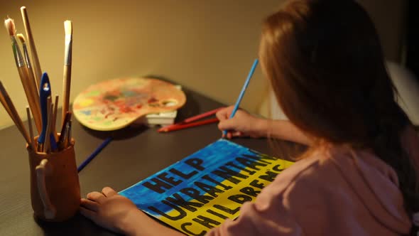 Shooting Over Shoulder of Cute Little Girl Painting Help Ukrainian Children Placard Sitting at Table alt
