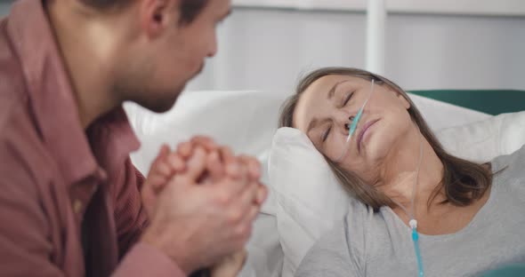 Close Up of Young Man Holding Hand of Sick Mother Sleeping in Hospital Bed alt
