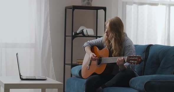Young Female Musician Is Playing Guitar Sitting at Home and Viewing Tutorial Video on Laptop alt