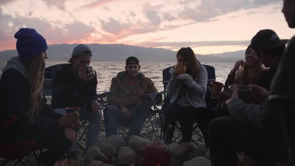 Group of friends drinking from mugs around campfire during sunset alt