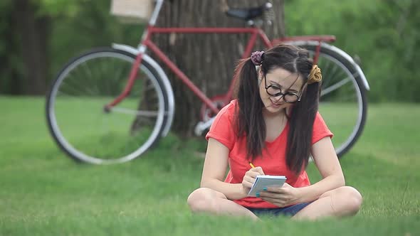 Young woman in red shirt with notebook sits on green grass with bike on background
