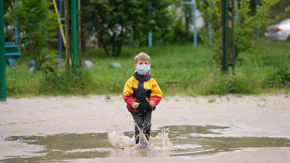 A small boy in a medical mask and rubber boots jumps through puddles on a spring day