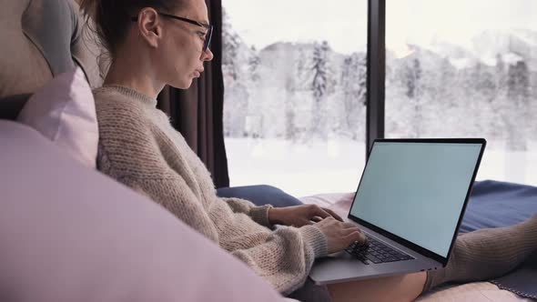 A Woman Freelancer with Glasses Sits on a Bed in a Country House with Panoramic Windows Behind Which alt