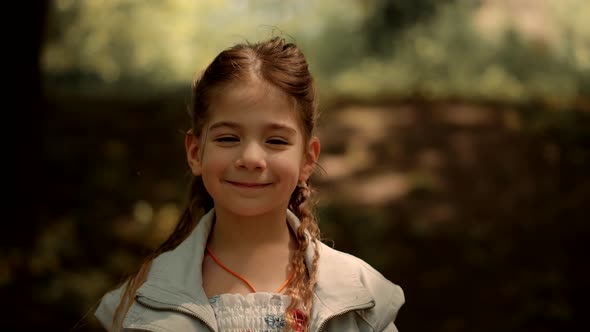 Cute Little Girl Portrait Smiles On Nature Looking At Camera On Botanical Garden. alt