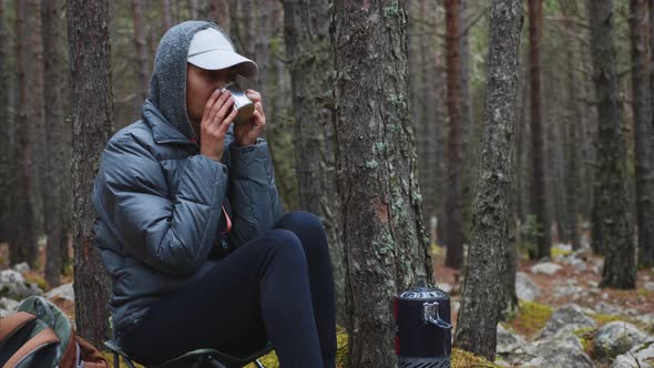 Woman Hiker in Jacket Drinks Hot Tea in Forest Camping Sitting on Chair