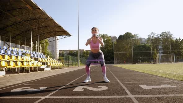 Slender Girl Squats with an Elastic Band and Jumps Up at the Stadium in Summer. alt