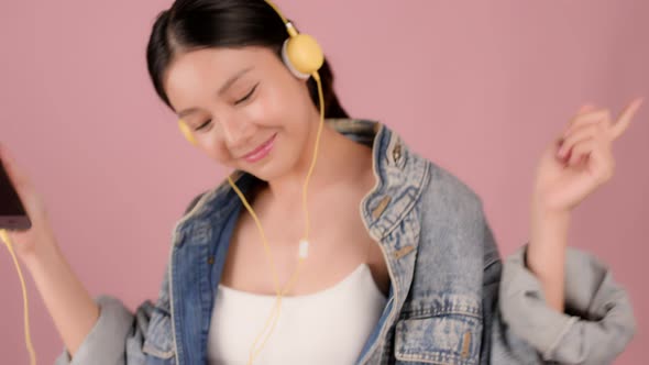 Happy pretty Asian teenager girl dancing and listening music by smartphone on pink background. alt