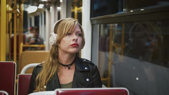 Female Hipster in Headphones is Riding a Tram Looking Out the Window and Listening to Music alt
