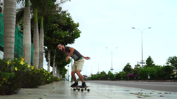 Millennial guy with man bun and beard skateboarding on empty sidewalk. Slow mo alt