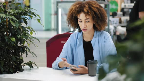 African American Girl Woman Sitting in Cafe with Coffee Looking at Mobile Phone Receiving Message alt