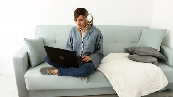 Attractive european blonde woman in headphones listening music and dancing sitting on comfy sofa