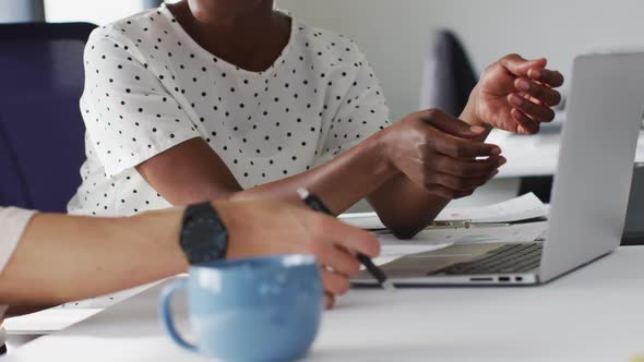 Midsectin of two diverse female colleagues looking at laptop and discussing in office alt