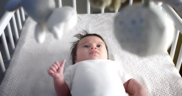 Newborn Baby in a White Bodysuit is Looking at a Baby Mobile in a Child Crib