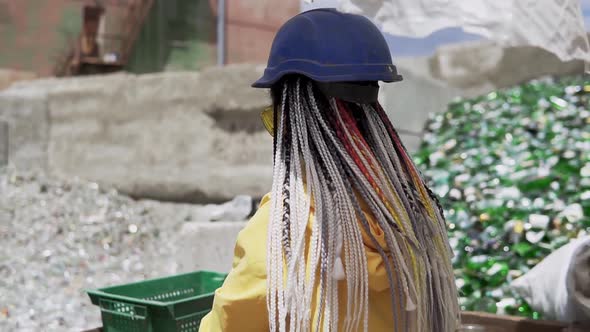 Young Woman in Hard Hat Standing Against the Pile of Broken Glass Used Bottles Next to the Wall alt